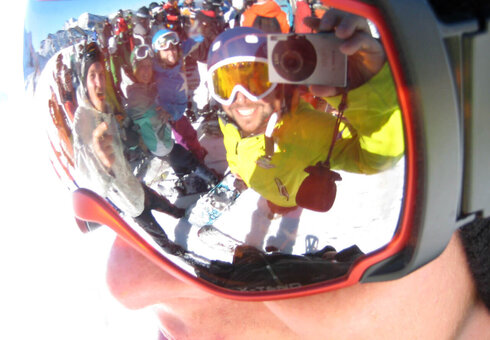 In einer Skibrille spiegelt sich eine Gruppe von Skifahrern, die ein Selfie macht